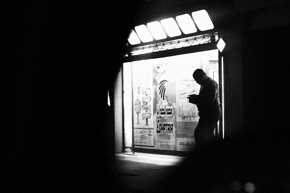 A contemplative silhouette captured in front of an EgyptAir sign, in a black and white street scene.
