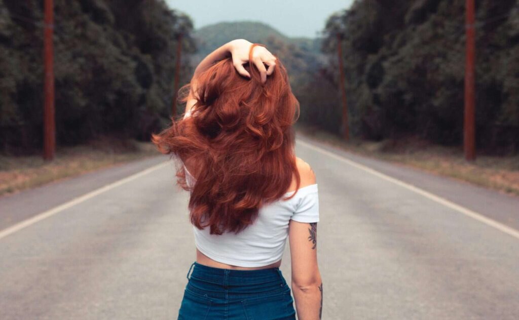 Red-haired woman in jean shorts walking down a forest road, showcasing style and freedom.