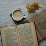 Warm overhead view of coffee, an open book, and yellow flowers on a fabric table.