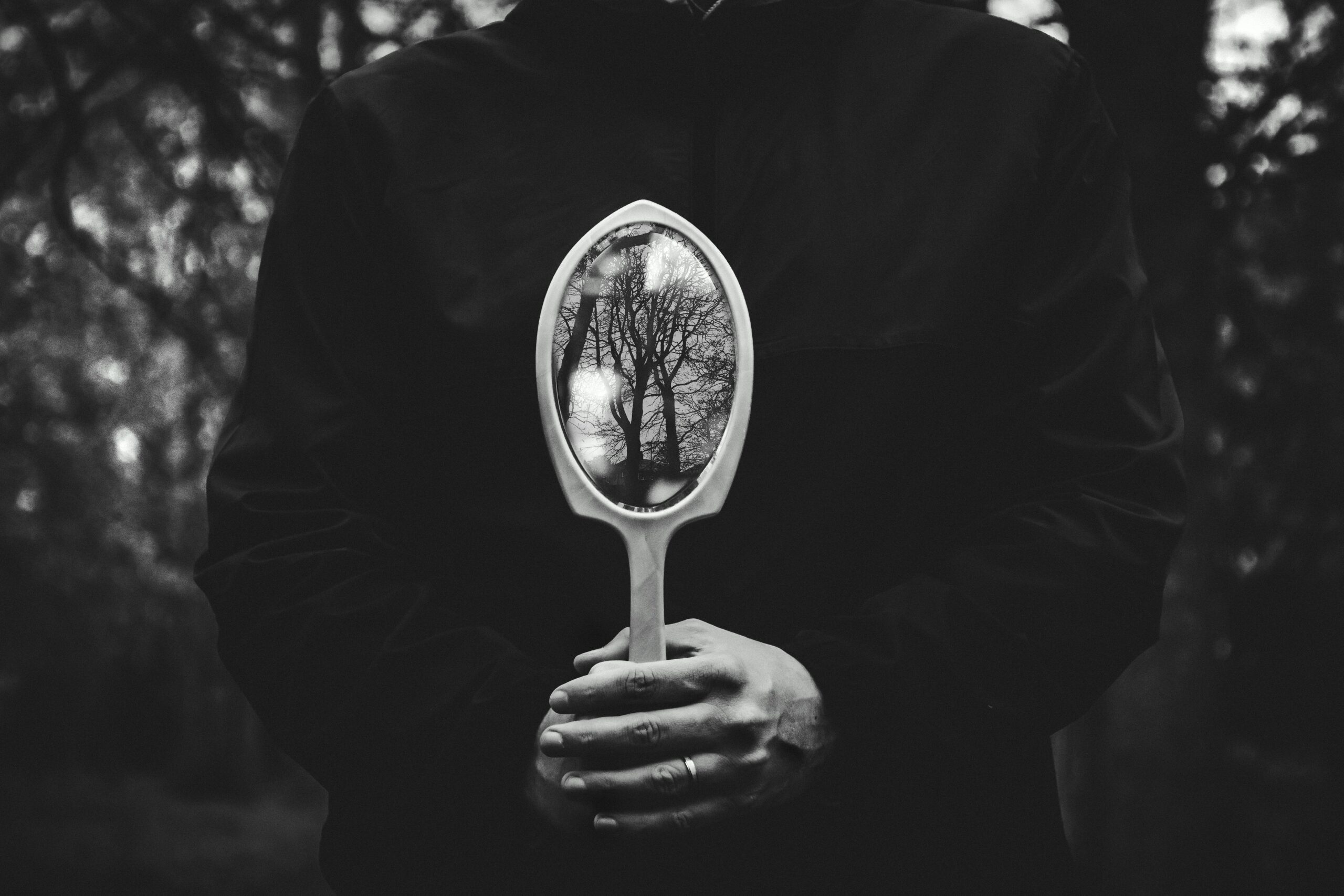 A person holding a mirror reflecting bare trees in a black and white setting.